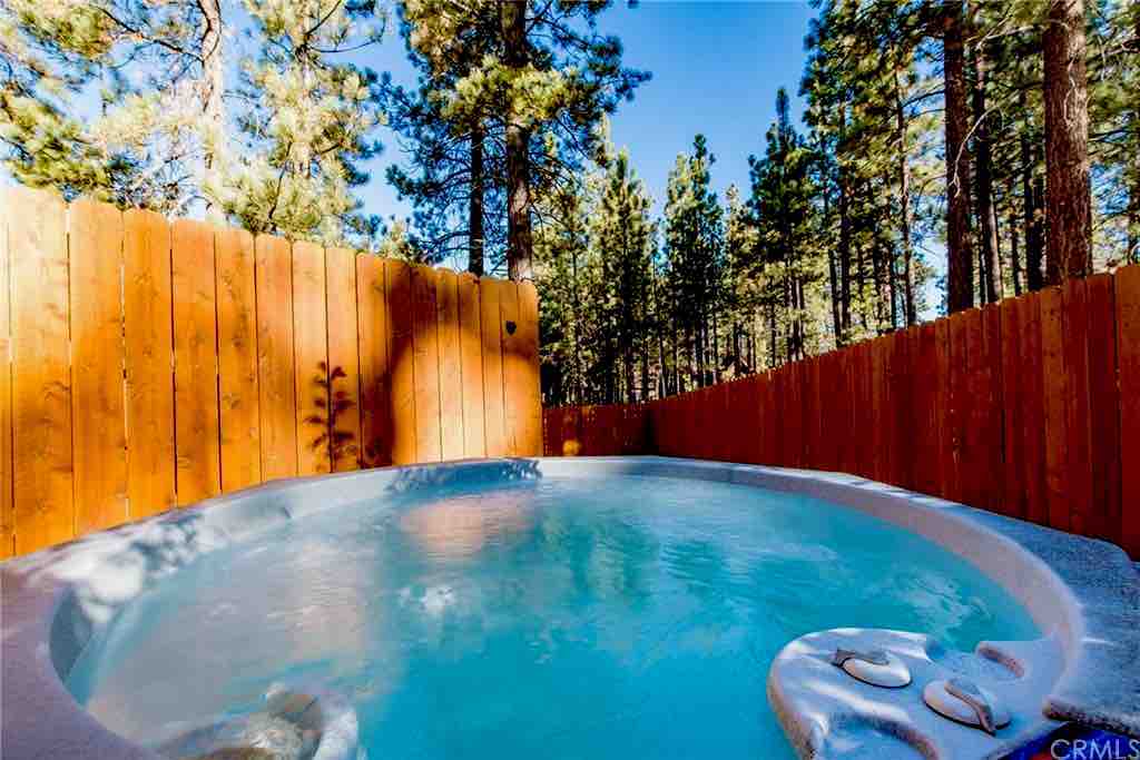A private jacuzzi is surrounded by fencing, offering a serene view of tall trees and clear skies. Water gently ripples, highlighting the inviting space designed for relaxation in a tranquil outdoor setting.