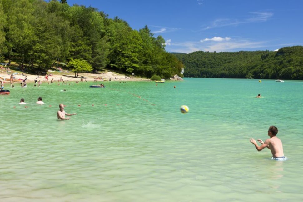 A tranquil beach scene at the Lac de Vouglans is depicted, featuring clear turquoise water and lush green trees lining the shore. Several individuals can be seen engaging in recreational activities, with some enjoying the water and others relaxing on the sandy beach.