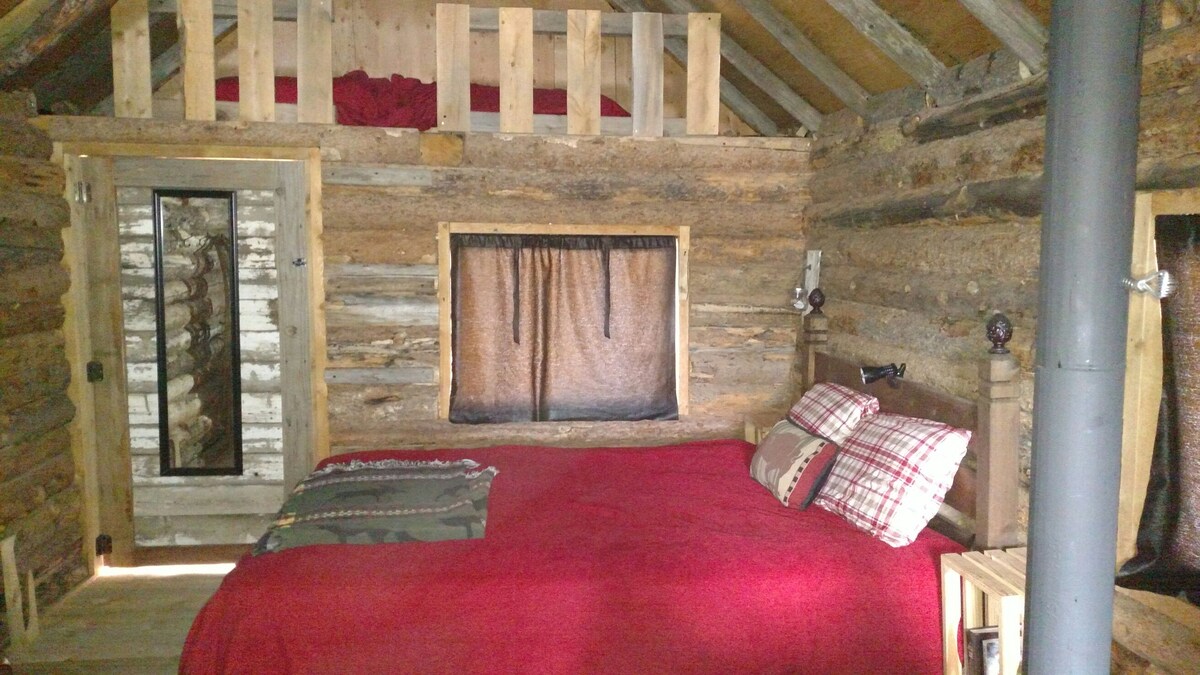 A cozy interior of a rustic log cabin features a spacious bed covered with a deep red blanket. Plaid accent pillows are placed at the head of the bed. A large window with dark fabric curtains provides natural light, while a wooden door and loft area are visible in the background.