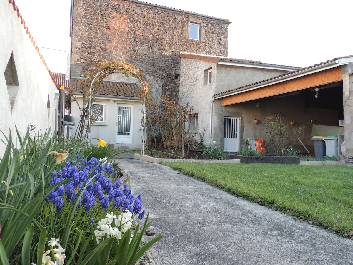 A stone house with a rustic façade stands in a tranquil garden setting, featuring a pathway lined with blooming flowers. The yard is complemented by a well-maintained lawn, with visible outdoor storage and subtle landscaping details enhancing the peaceful environment.