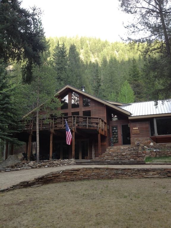 The exterior of a wooden home is framed by towering trees, showcasing a spacious balcony with rustic railings. A stone pathway leads to the entryway, complemented by a manicured lawn. An American flag is displayed prominently at the front, adding a welcoming touch.