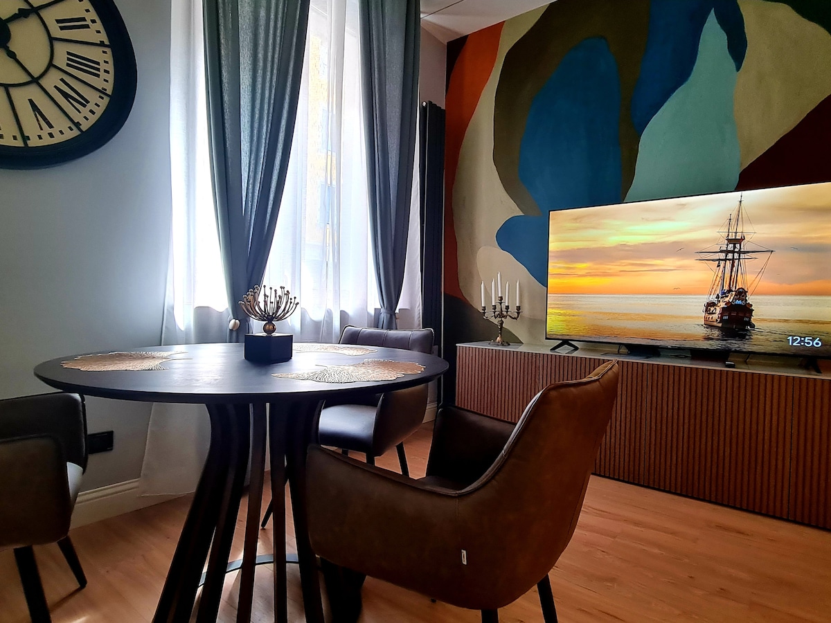 A modern living area features a round black dining table with three upholstered chairs. A large TV displays a scenic image, and a colorful mural offers an artistic backdrop. Natural light streams through sheer curtains, enhancing the room's inviting ambiance.