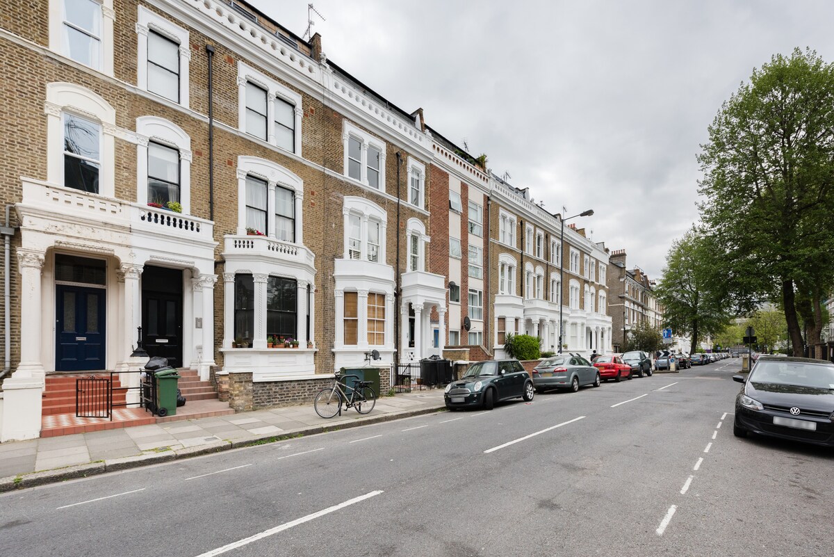 A row of traditional Victorian buildings lines the street, characterized by their distinct architectural details. Large windows are visible, with a mix of residential and parked vehicles along the road. Trees provide greenery, contributing to the urban atmosphere.