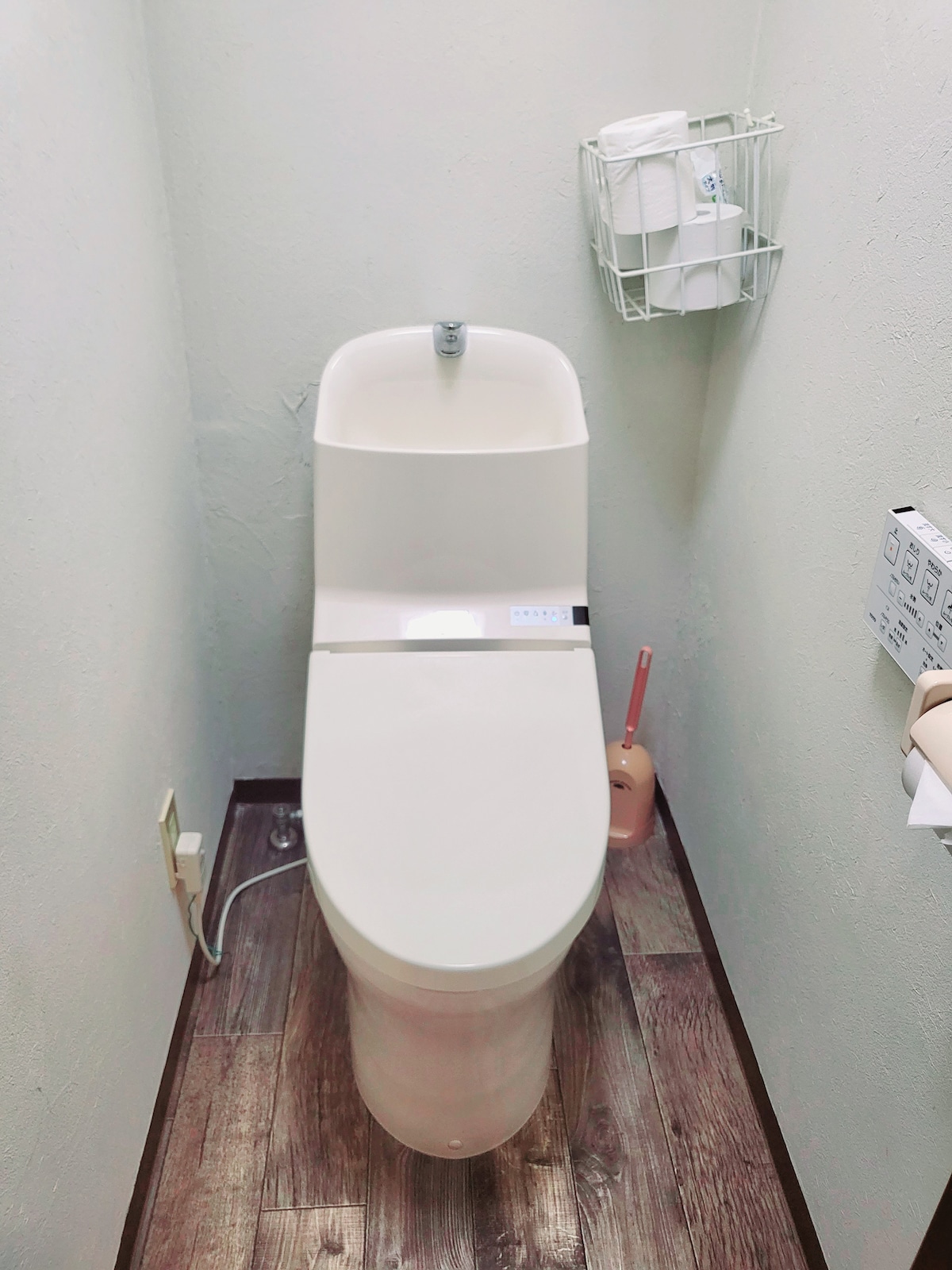 A modern toilet is positioned in a well-lit, compact space. A wire basket holds neatly arranged toiletries nearby, while the wall features essential amenities. The floor is covered with textured tiles, contributing to the room's functional design.
