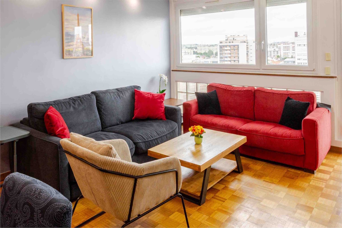 The living room features two sofas and an armchair arranged around a coffee table. A wall-mounted artwork is visible, and natural light enters through large windows, providing a view of the surroundings. The wooden floor adds warmth to the space.