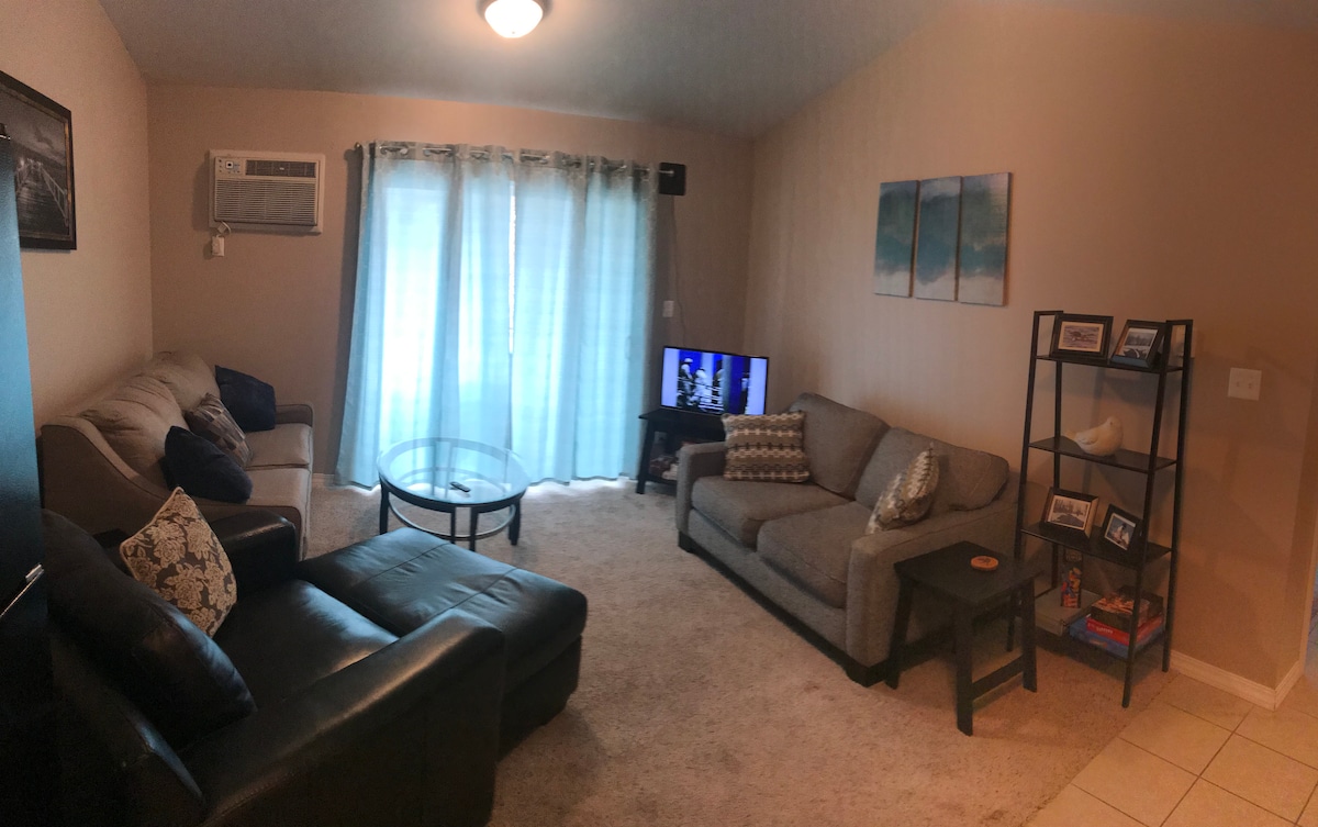 A cozy living room showcases two sofas and a stylish armchair positioned around a glass coffee table. Natural light filters through sheer curtains, highlighting the neutral tones of the decor. A television is placed on a functional shelving unit that also displays decorative items.