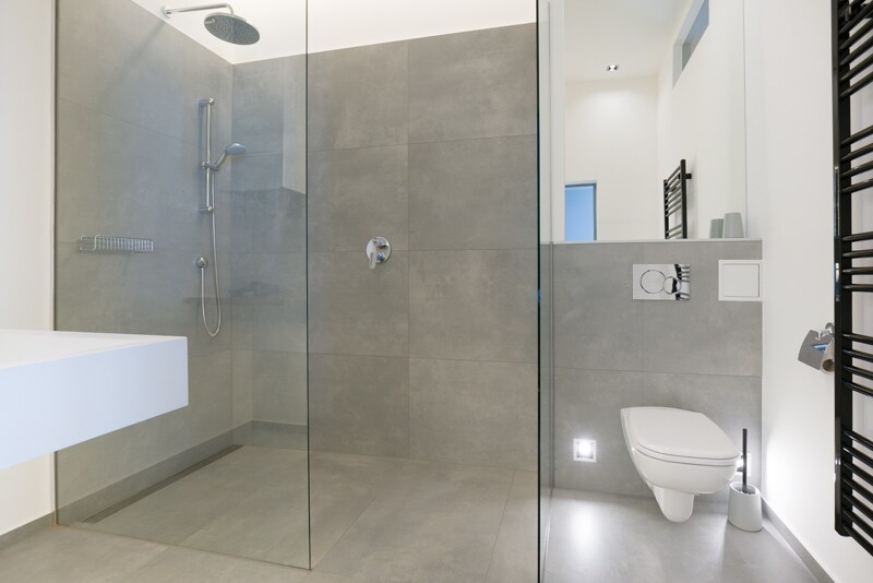 A modern and spacious bathroom features a glass-enclosed shower with rainfall and handheld shower heads. A sleek floating vanity with a minimalist design is complemented by a wall-mounted toilet. Neutral tones dominate the palette, ensuring a clean and contemporary aesthetic.