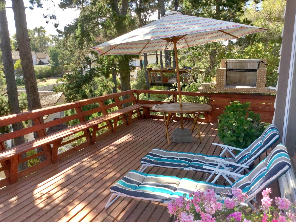 A wooden deck is seen adorned with two lounge chairs featuring striped cushions. A large umbrella provides shade over a round table, surrounded by greenery and colorful flowers. A barbecue grill is visible in the background, contributing to the outdoor dining experience.