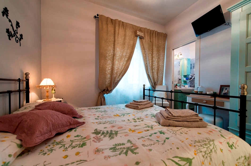 A comfortable bedroom features a double bed with floral bedding, complemented by two decorative pillows. Soft curtains frame the window, allowing natural light to fill the room. A small lamp provides additional illumination beside the bed, with a mirror and vanity area visible in the background.