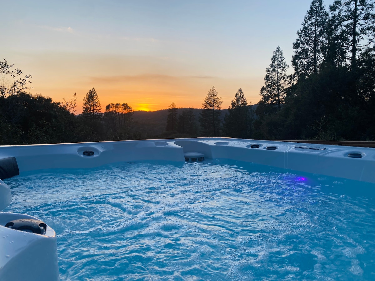 A spacious hot tub is filled with bubbling water, set against a stunning sunset backdrop. Trees and rolling hills can be seen in the distance, with soft light illuminating the scene, creating a tranquil atmosphere for relaxation.