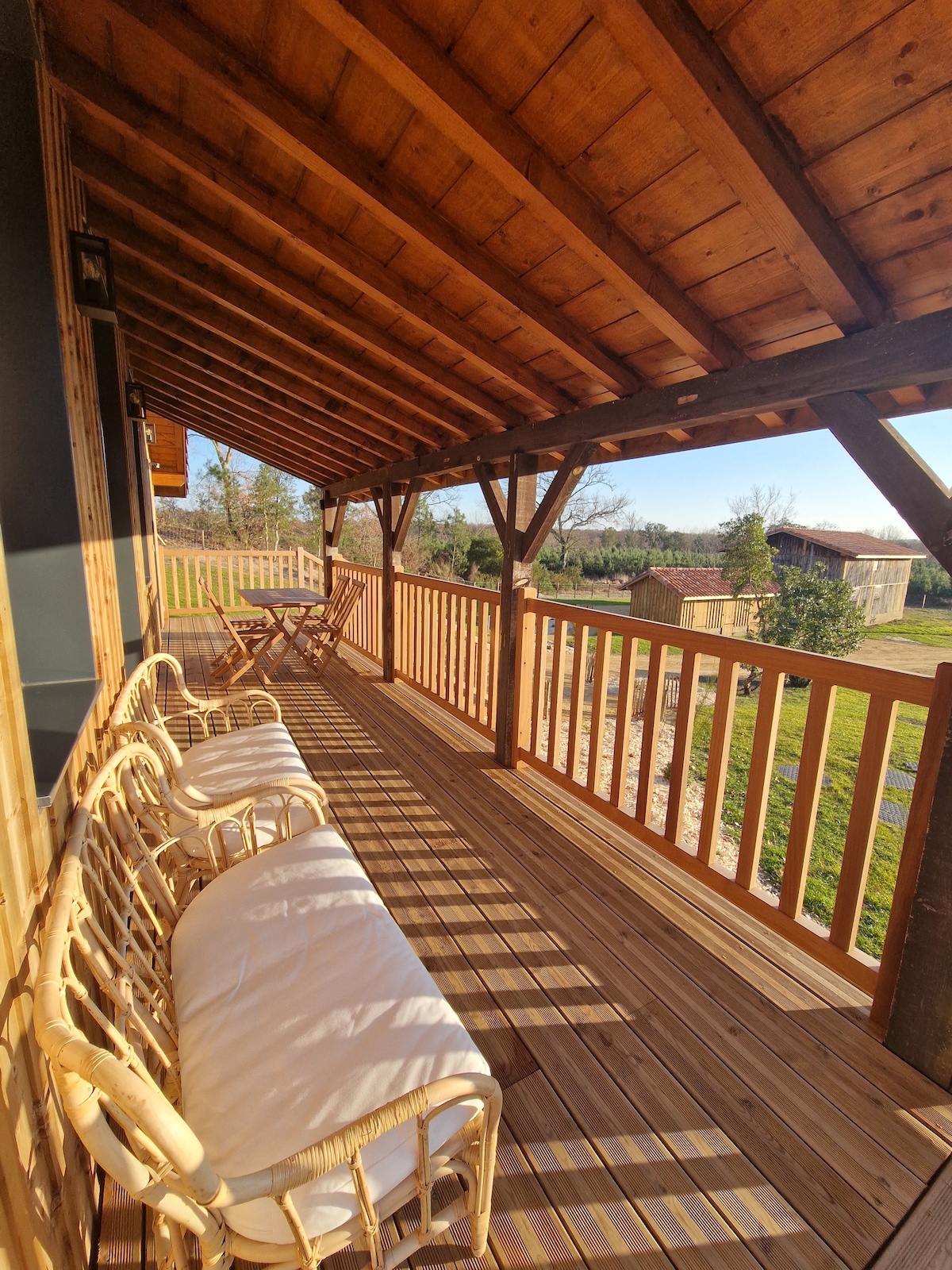 A spacious wooden terrace is presented, featuring a covered area supported by wooden beams. Two wicker chairs with soft cushions are arranged beside a small table, overlooking green surroundings and allowing for a serene outdoor seating experience.
