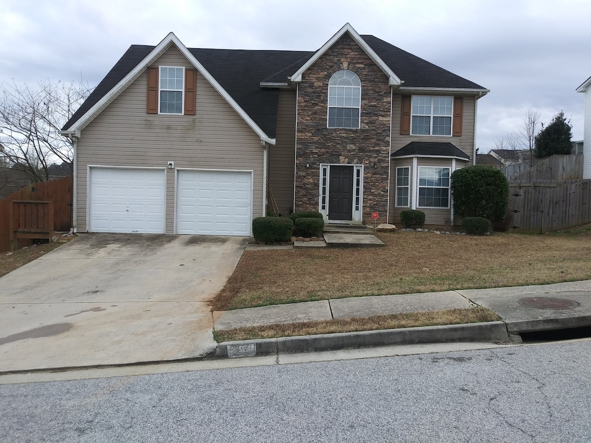 The exterior of a two-story home is displayed, featuring a combination of stone and siding materials. A two-car garage is located on the left, with a concrete driveway leading to the front entrance. The yard is well-maintained, with shrubs flanking the pathway.