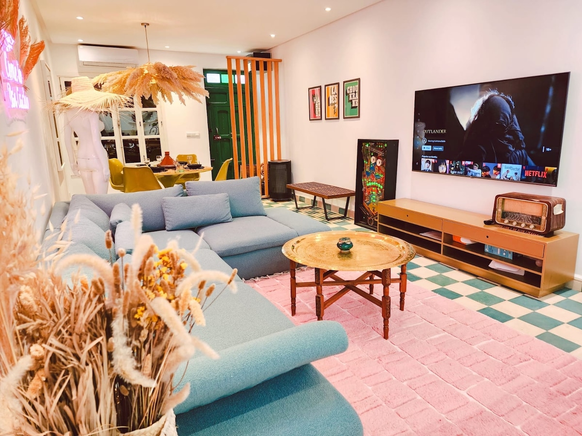 A spacious living area is highlighted by a large sectional sofa in a soft blue fabric. A vintage-style coffee table occupies the center, surrounded by decorative elements. A flat-screen TV on the wall displays entertainment options. Colorful artwork adorns the walls, enhancing the design.