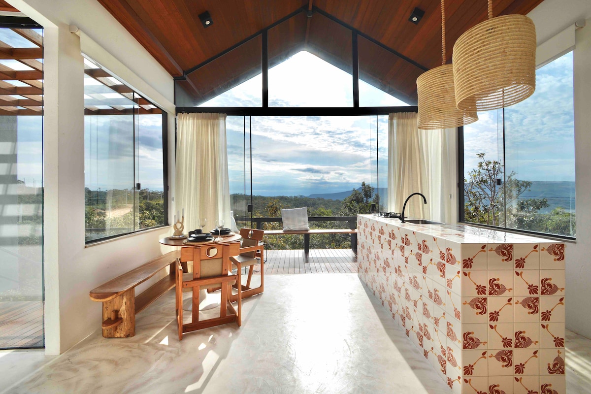 A bright living area integrates with the outdoors, featuring large glass windows that frame a mountain view. A compact kitchen island with unique tile design is complemented by a wooden dining table and benches, creating a harmonious space for relaxation and dining.