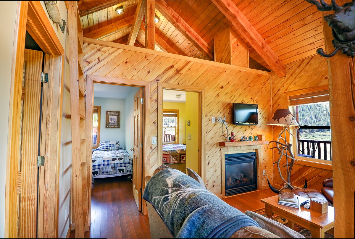 The interior of the cabin features wooden walls and ceilings, enhancing the rustic feel. A comfortable seating area is visible, complemented by a propane fireplace. A doorway leads to a bedroom with two single beds, while a Smart TV is mounted on the wall, providing entertainment options.