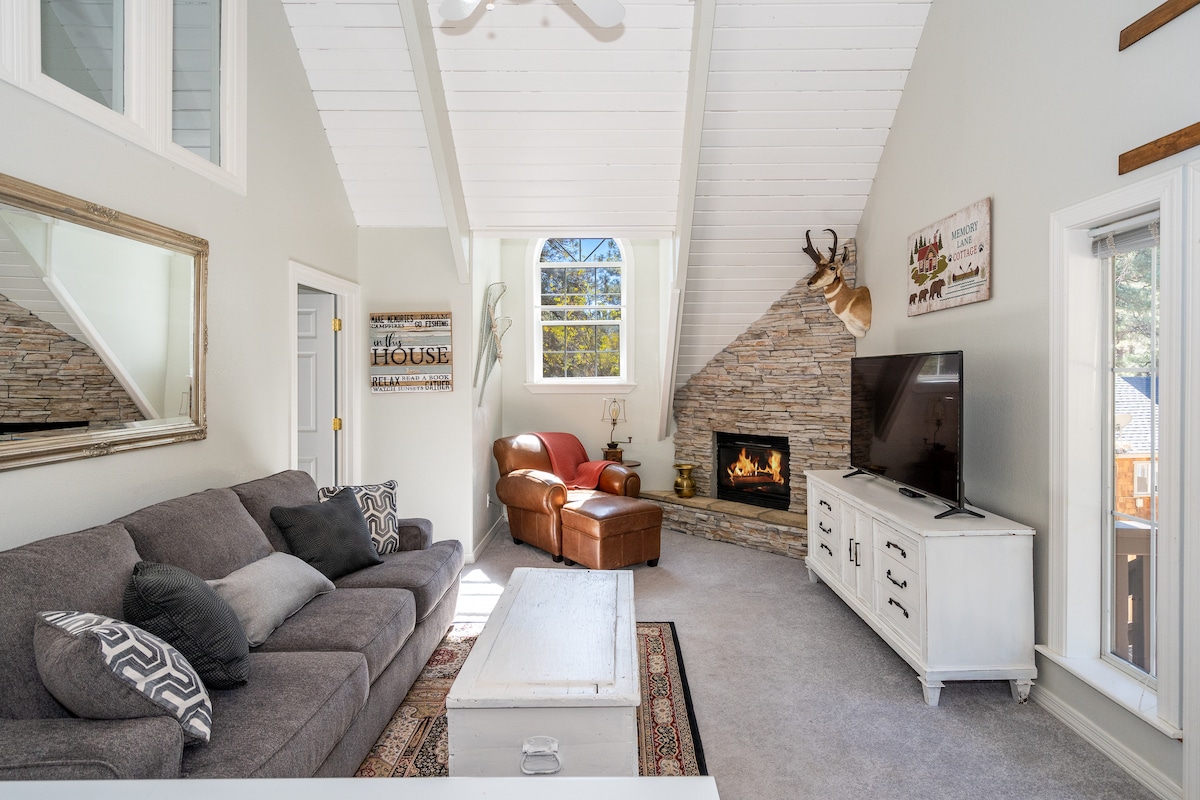The living area features a light-colored sofa arranged across from a stone fireplace. Large windows allow natural light to illuminate the space, while a neutral-toned area rug adds warmth beneath a coffee table. A flat-screen television is mounted on a nearby wall.