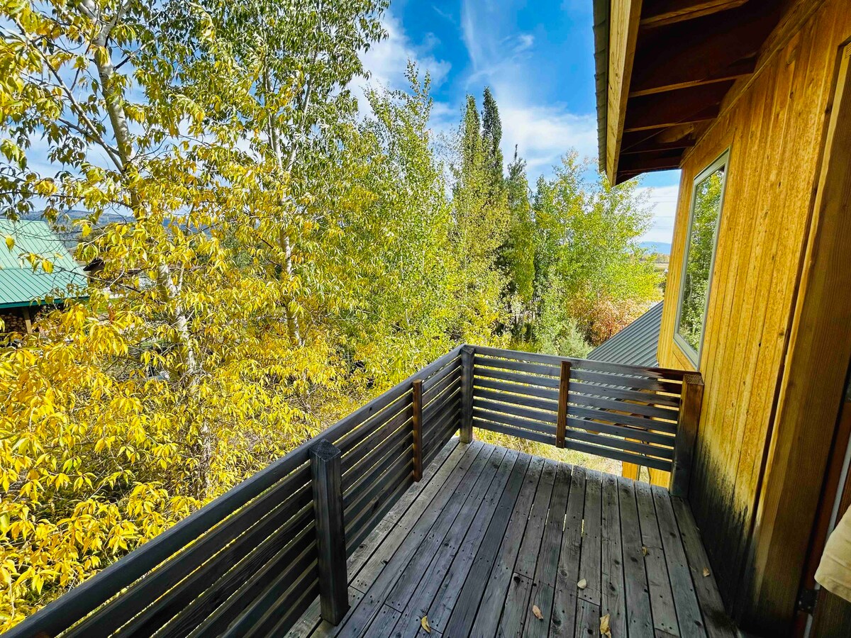 A wooden balcony features slatted railings and overlooks a vibrant landscape of golden-leaved trees. The sky above displays a mix of blue and white clouds, creating a serene outdoor setting. The balcony is attached to a rustic wooden structure, highlighting natural elements.