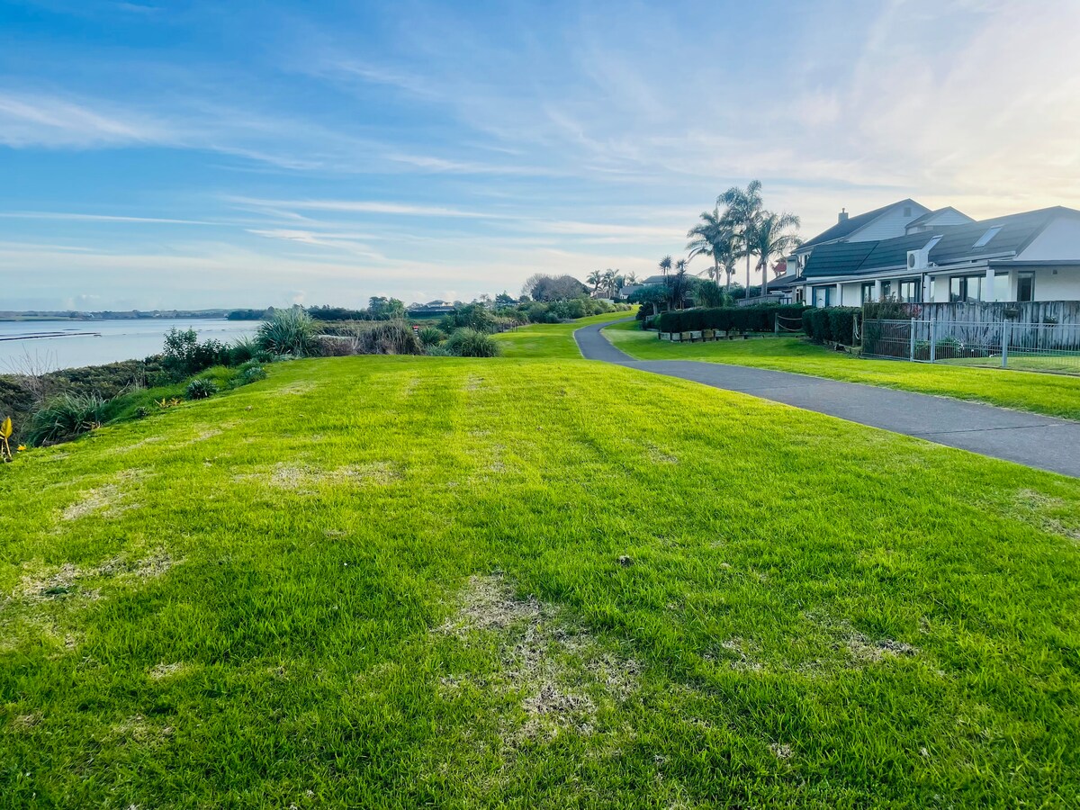 A green, expansive lawn extends towards a walking path beside a freshwater stream. In the background, a residential building is visible, complemented by palm trees. The setting is tranquil, with a clear sky and soft sunlight illuminating the area.