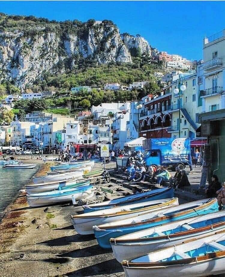 Colorful boats are docked along a rocky shoreline. A vibrant village is set against a backdrop of steep cliffs, with houses nestled into the hillside. Clear blue skies enhance the lively atmosphere, while people are seen enjoying the marina.