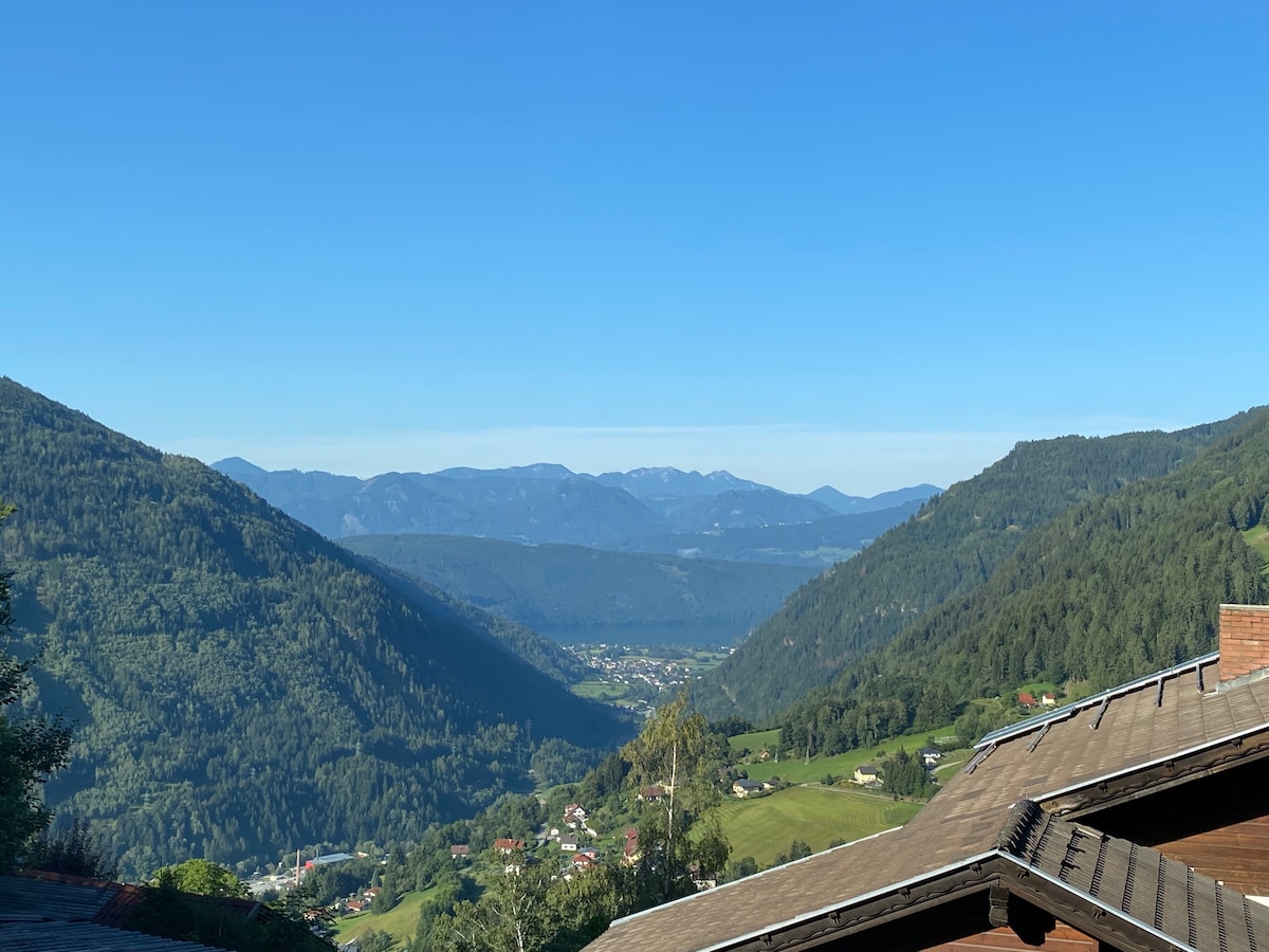 A panoramic view across the Nockbergen mountains reveals a vast landscape of rolling hills and valleys under a clear blue sky. Lush greenery extends into the distance, with hints of small villages nestled in the foothills. The natural beauty invites exploration and relaxation.