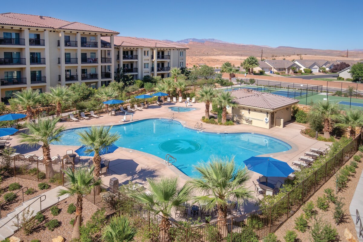 A spacious outdoor pool area is surrounded by palm trees and lounge chairs, offering ample seating. The pool features clear blue water, and an adjacent tennis court is visible in the background. A three-story building is positioned to the left, blending harmoniously with the landscape.
