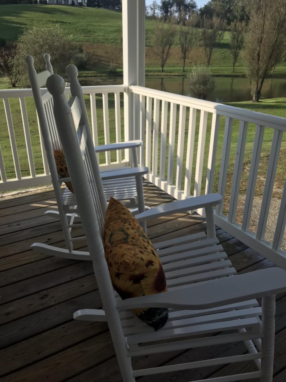 A private porch features two white rocking chairs positioned beside a colorful pillow. The serene landscape of green hills and a pond is visible in the background, offering a peaceful outdoor setting.