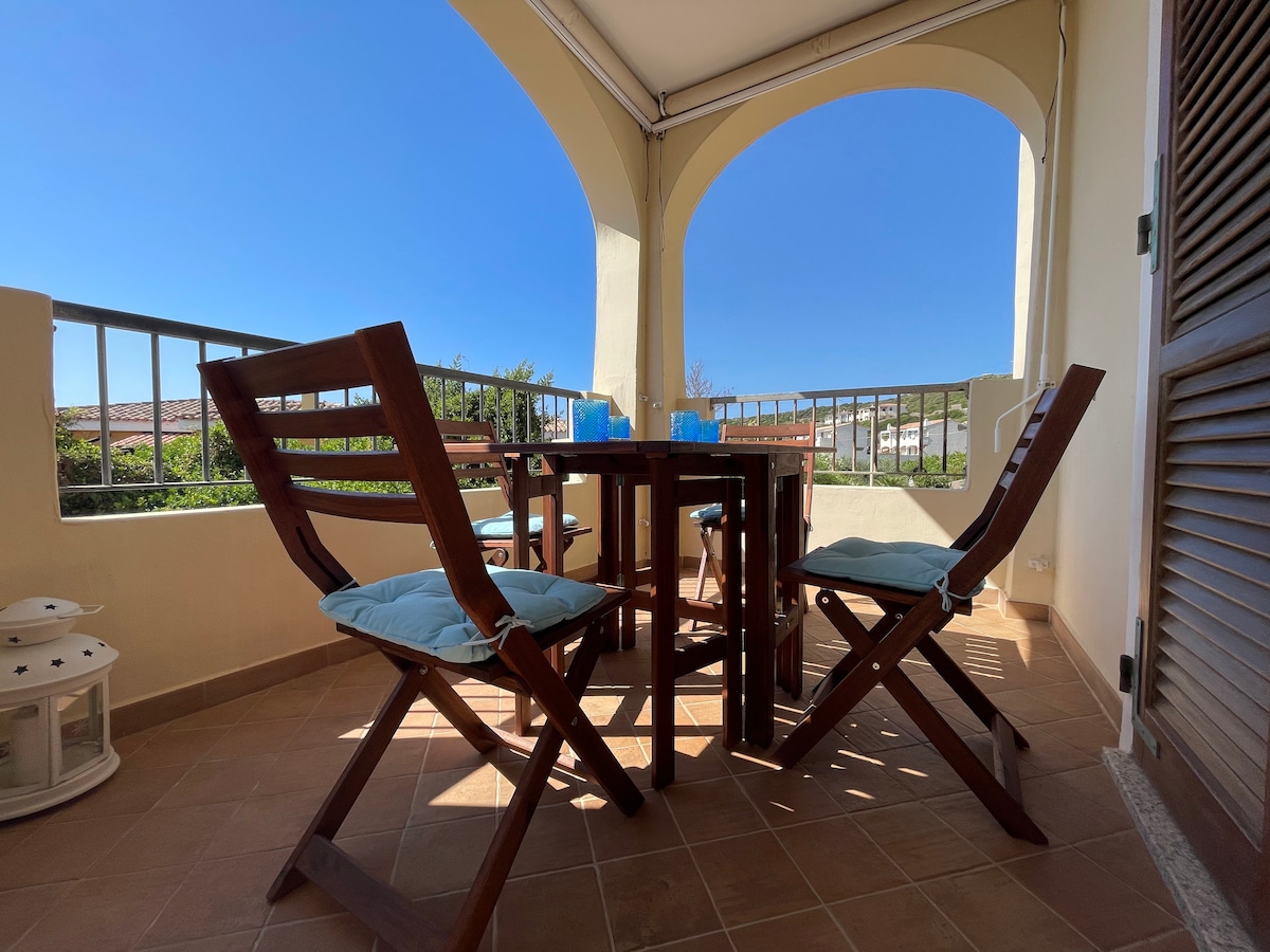 A spacious terrace is depicted, featuring a wooden table surrounded by foldable chairs with blue cushions. The area is enclosed by a railing, with clear skies and greenery visible in the background, providing a pleasant setting for gatherings.