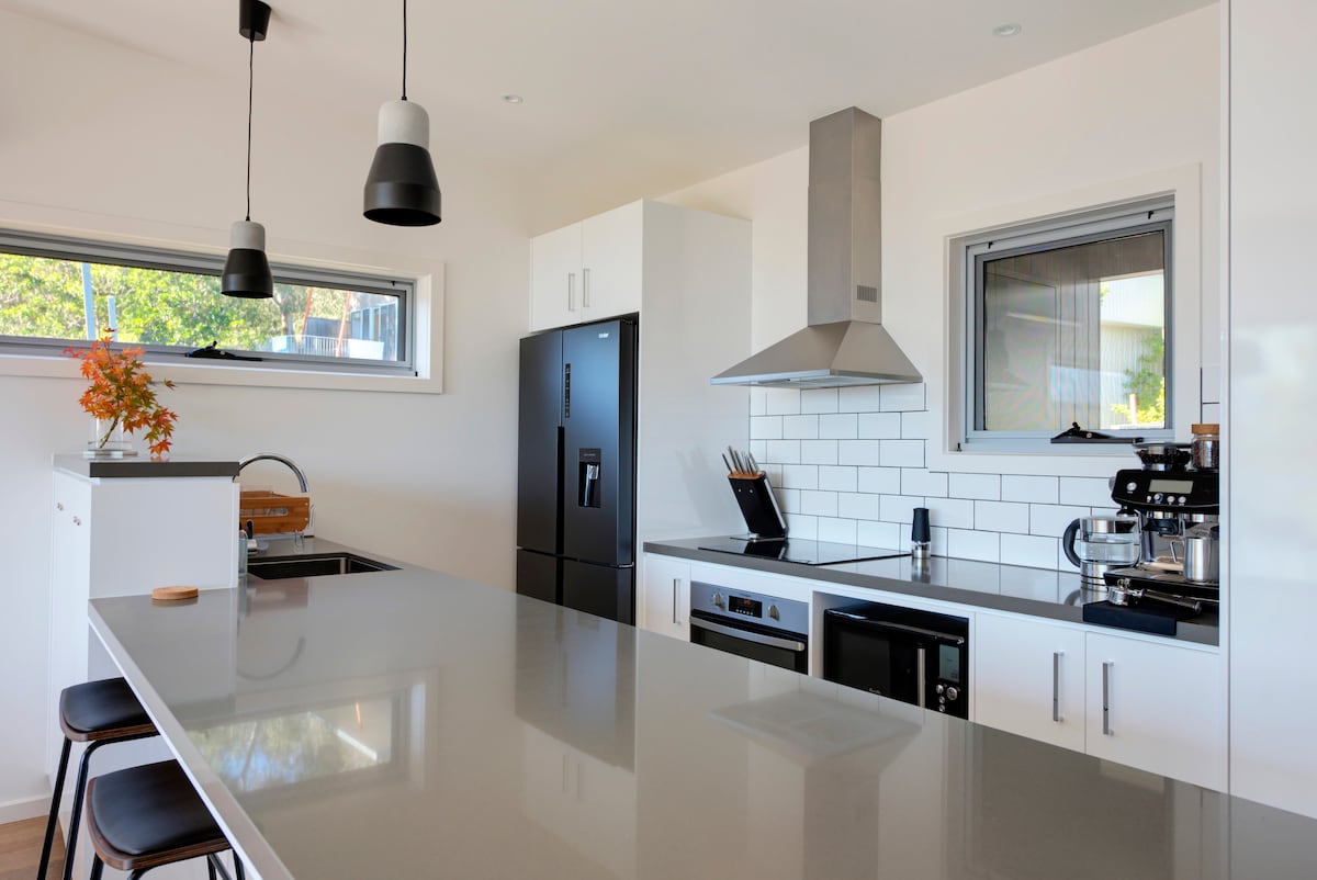 The modern kitchen features sleek cabinetry and a spacious island with seating. Stainless steel appliances, including a refrigerator and oven, are integrated into the design. Natural light pours in through the windows, enhancing the inviting atmosphere of the workspace.