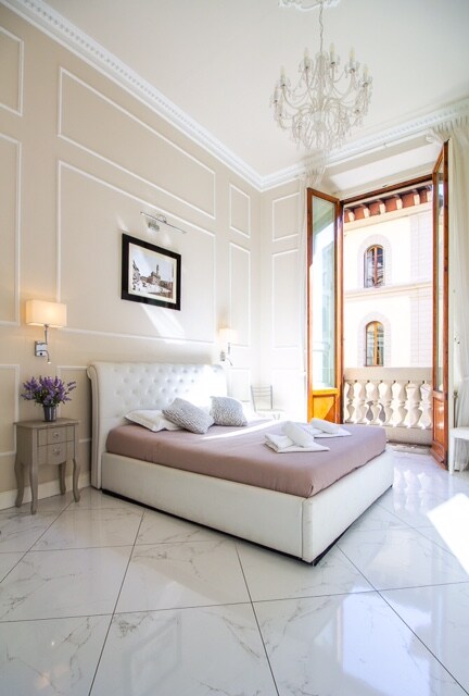 A light-filled bedroom features a spacious bed with neutral linens. A chandelier hangs from the ceiling, and natural light enters through an open balcony door. A small nightstand with a floral arrangement complements the elegant wall detailing and polished marble flooring.