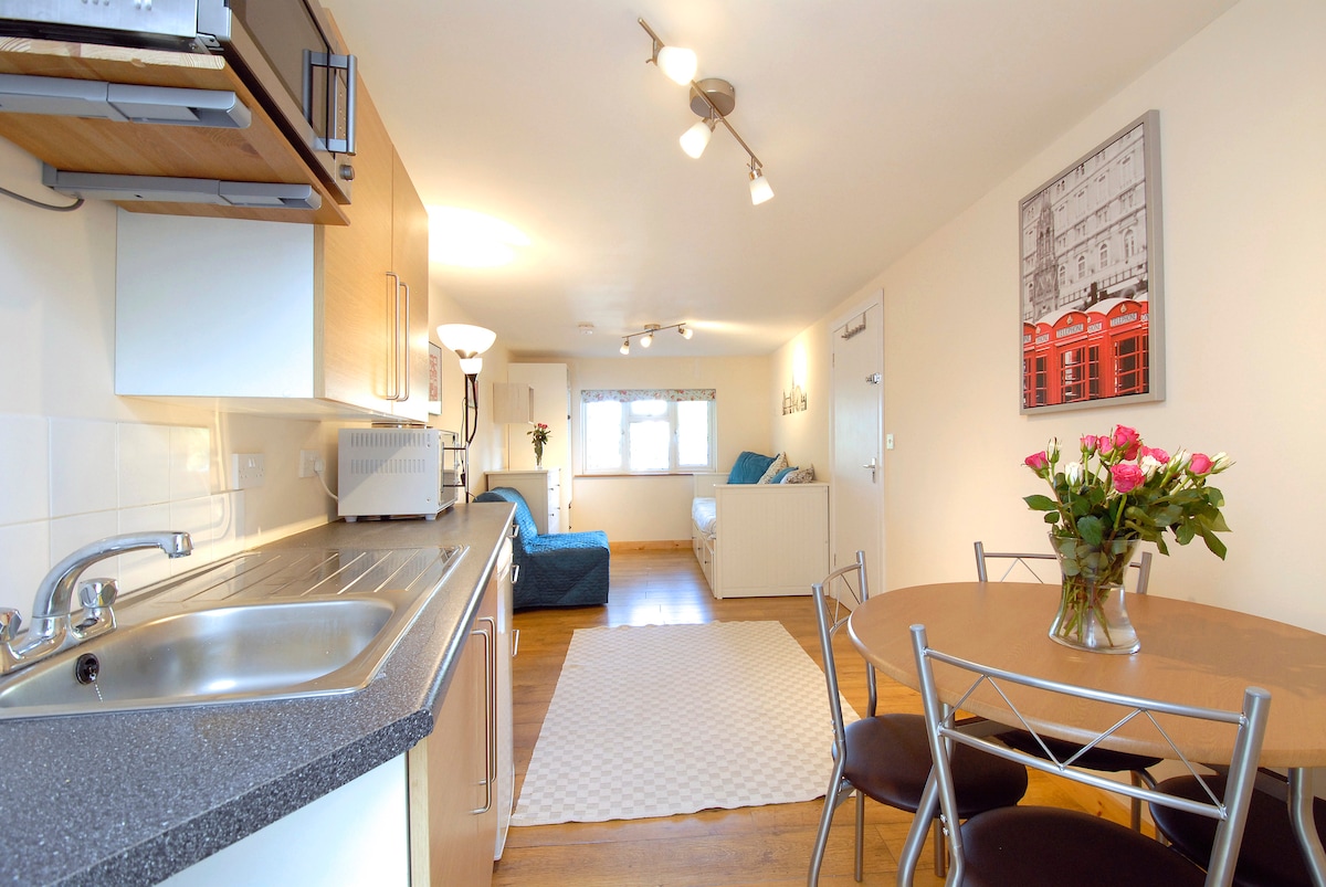 An open kitchen and dining area showcases a sleek countertop with a sink, microwave, and cooking appliances. A round dining table with four chairs is set beside a bouquet of flowers. Natural light filters through the window, illuminating the cozy sitting area in the background.