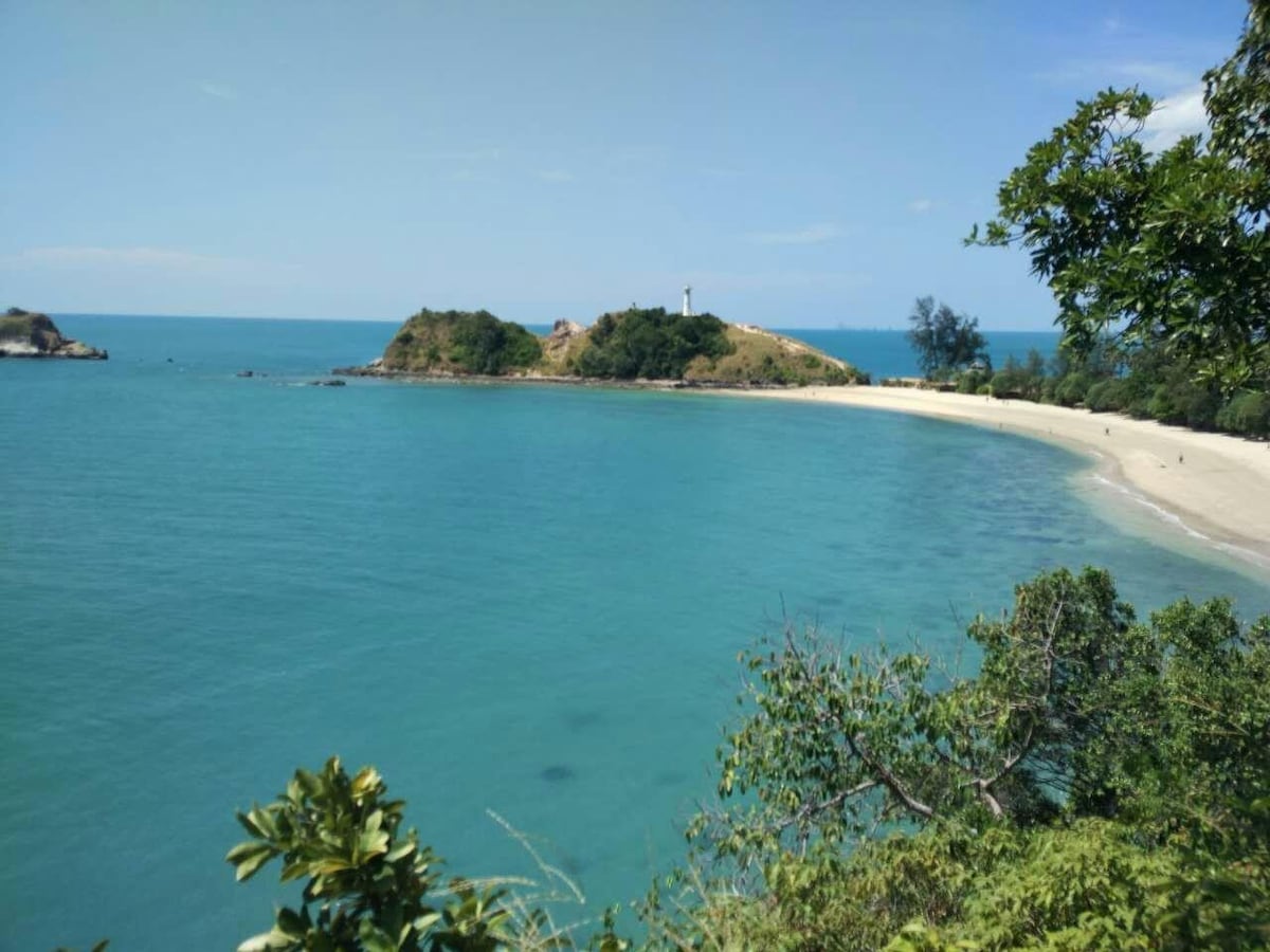 A serene coastal scene is depicted, featuring clear turquoise waters gently lapping the shore. Golden sandy beaches curve along the coastline, framed by lush greenery. In the distance, a small island with a lighthouse is visible, set against a backdrop of a bright blue sky.