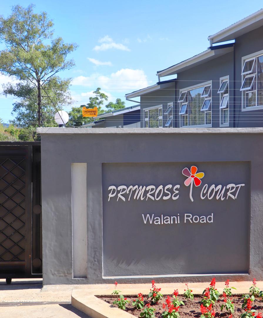 The entrance to Primrose Court is marked by a modern sign with vibrant lettering, set against a clean gray wall. A blossoming flower garden features red flowers, and the building displays large windows on both sides under a clear blue sky.