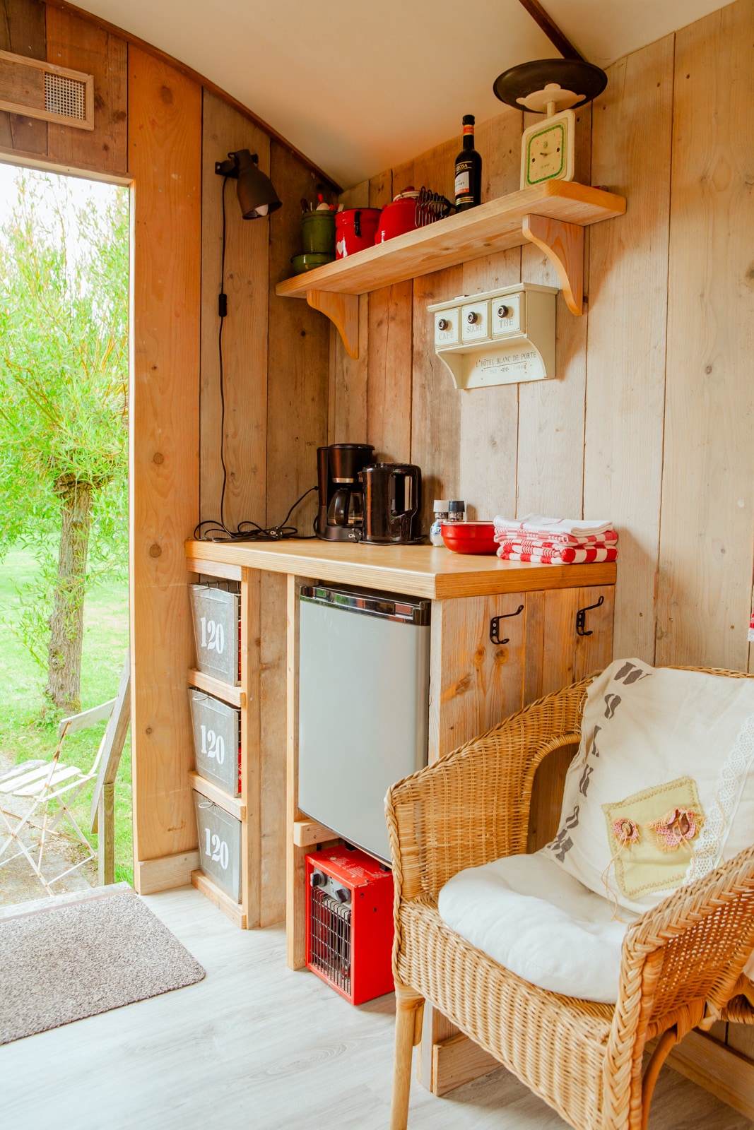 A compact kitchenette features wooden shelving with essential kitchen items, including a coffee maker and refrigerator. A wicker chair adds seating comfort, while sunlight filters in from the open door, illuminating the natural surroundings just outside.