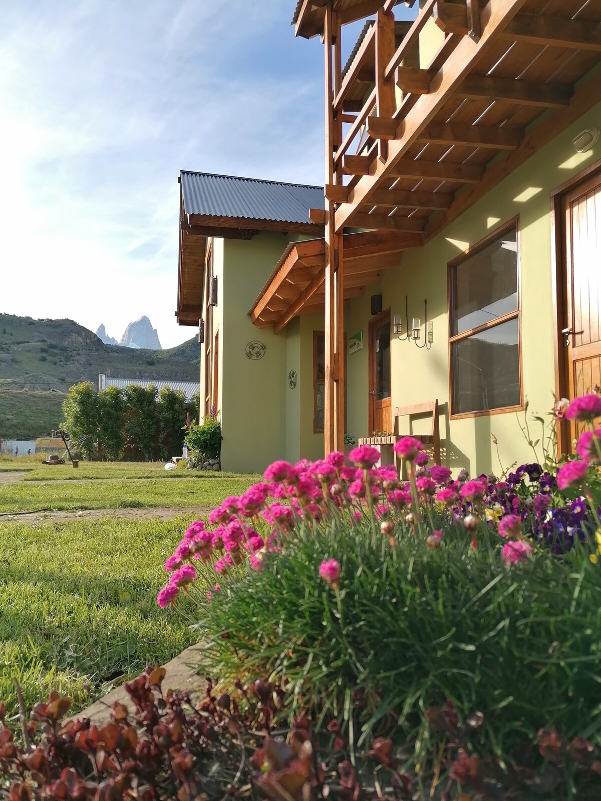 A charming exterior view of the accommodation is presented, showcasing a well-maintained garden with vibrant pink flowers and lush greenery. The building features wood accents and large windows, providing a bright and inviting atmosphere. Majestic mountain peaks are visible in the background.