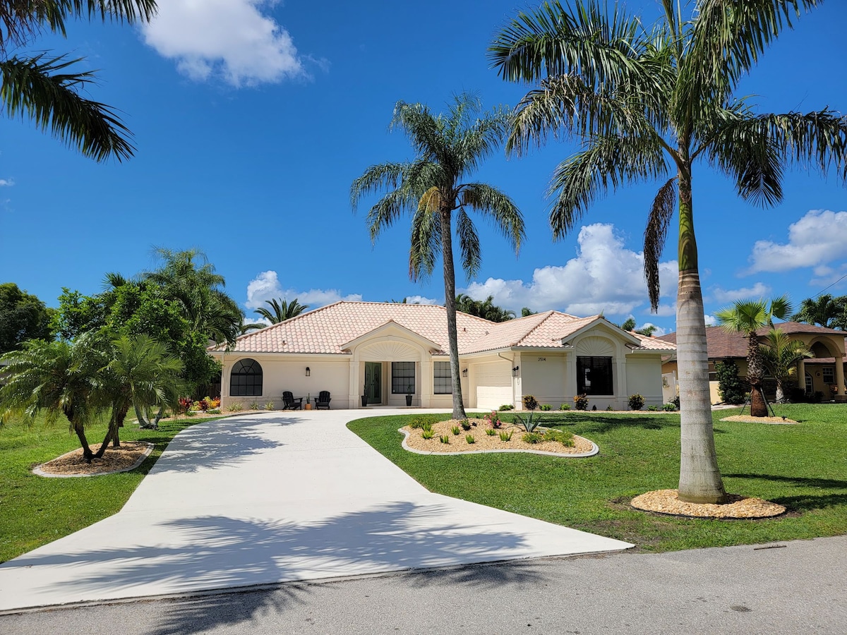 A single-story home is showcased with a light-colored exterior and a tiled roof. Lush greenery, including palm trees, surrounds the property. The driveway is smoothly paved, leading towards the entrance, with neatly arranged flower beds planted along the path.
