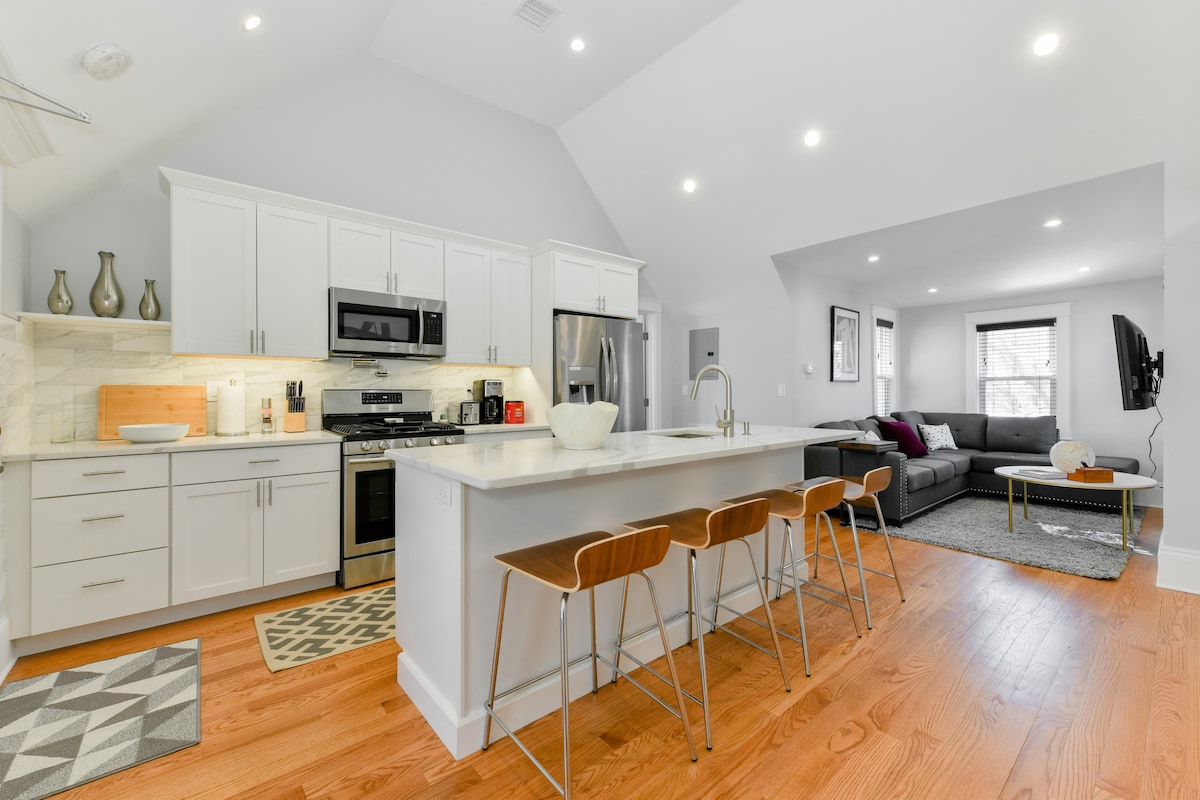 An open-concept kitchen and living area is presented, featuring white cabinetry and modern appliances. A large island with three bar stools creates a gathering space. The living room includes a comfortable sofa and a TV mounted on the wall, with natural light coming from nearby windows.