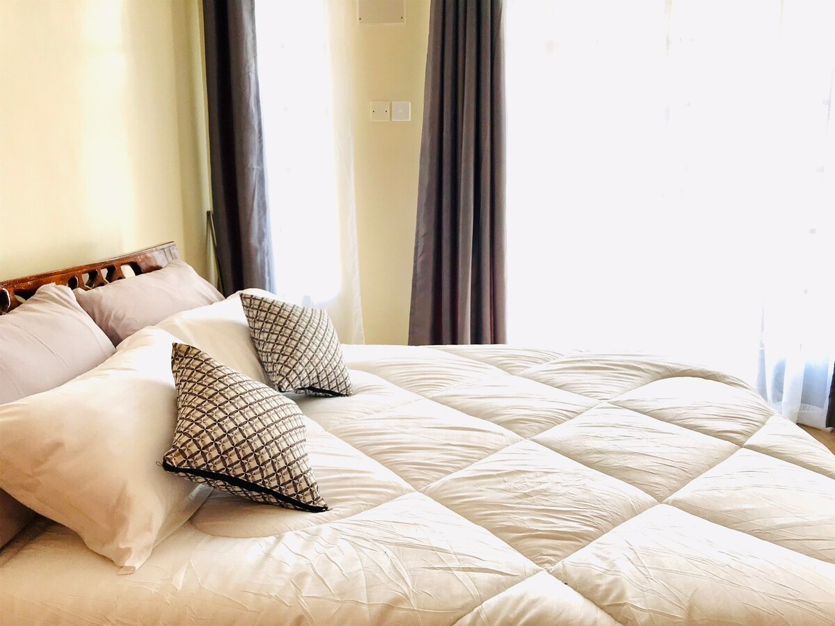 A cozy sleeping area features a spacious bed dressed in crisp white linens, complemented by two decorative pillows. Soft light enters through large windows, with sheer curtains providing a gentle filter. The serene color palette creates a relaxing ambiance.