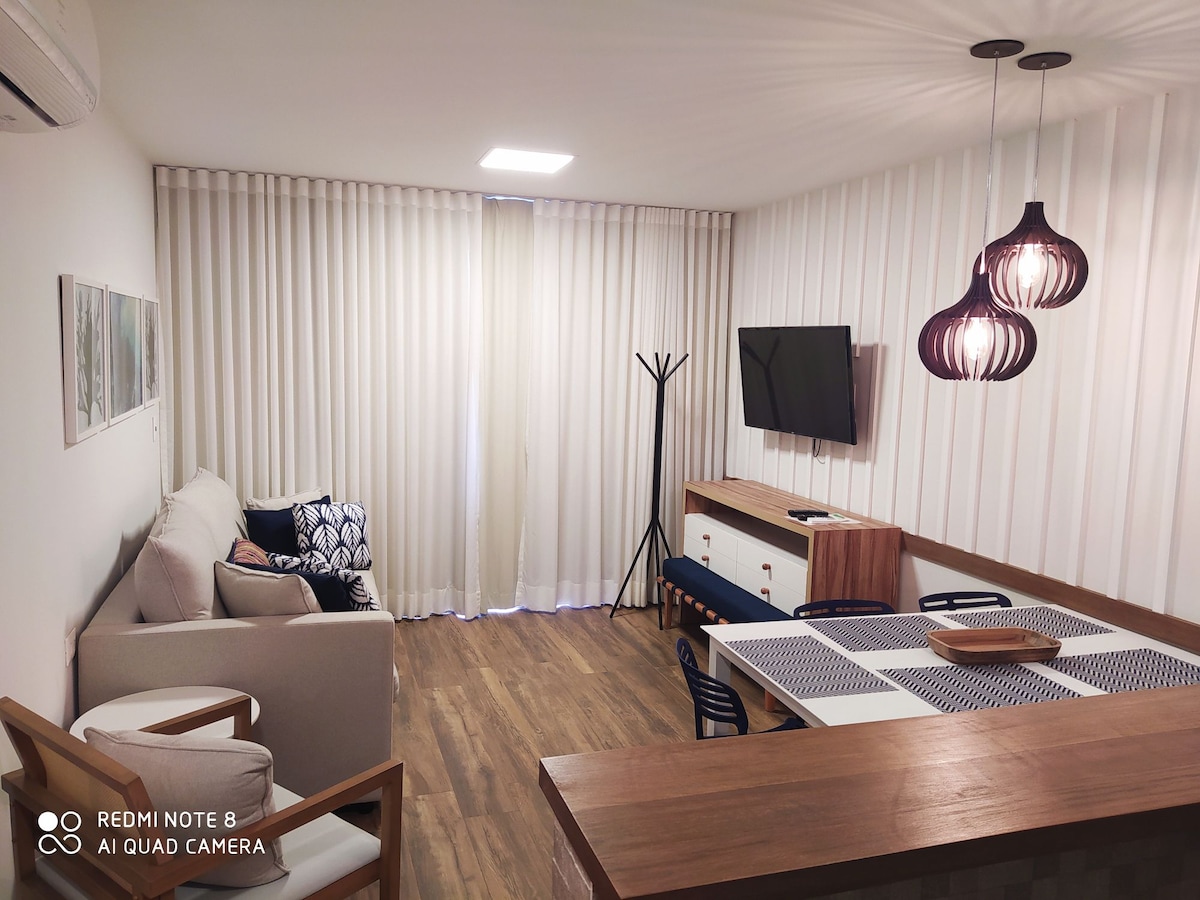 A cozy living area is presented with a light-colored sofa, a television mounted on the wall, and decorative lighting fixtures. Soft curtains drape over the window, while a dining table with wooden accents is visible, accompanied by stylish chairs and a modern sideboard.