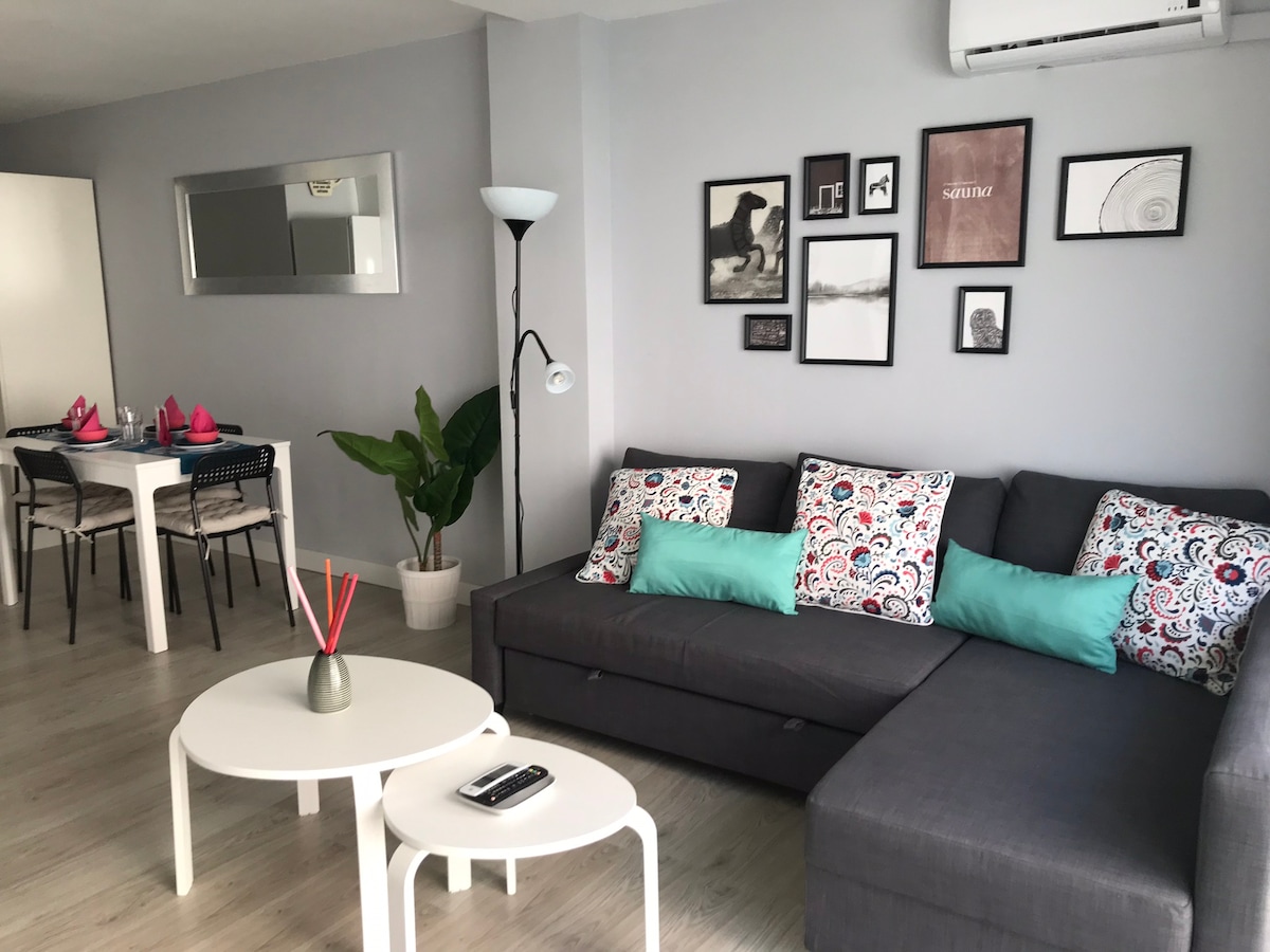 A modern living area is shown, featuring a dark gray sectional sofa adorned with decorative cushions. A white coffee table is centered in front of the sofa, while a dining table with black chairs is visible in the background. Wall art adds a decorative touch, and a green plant offers a natural element.