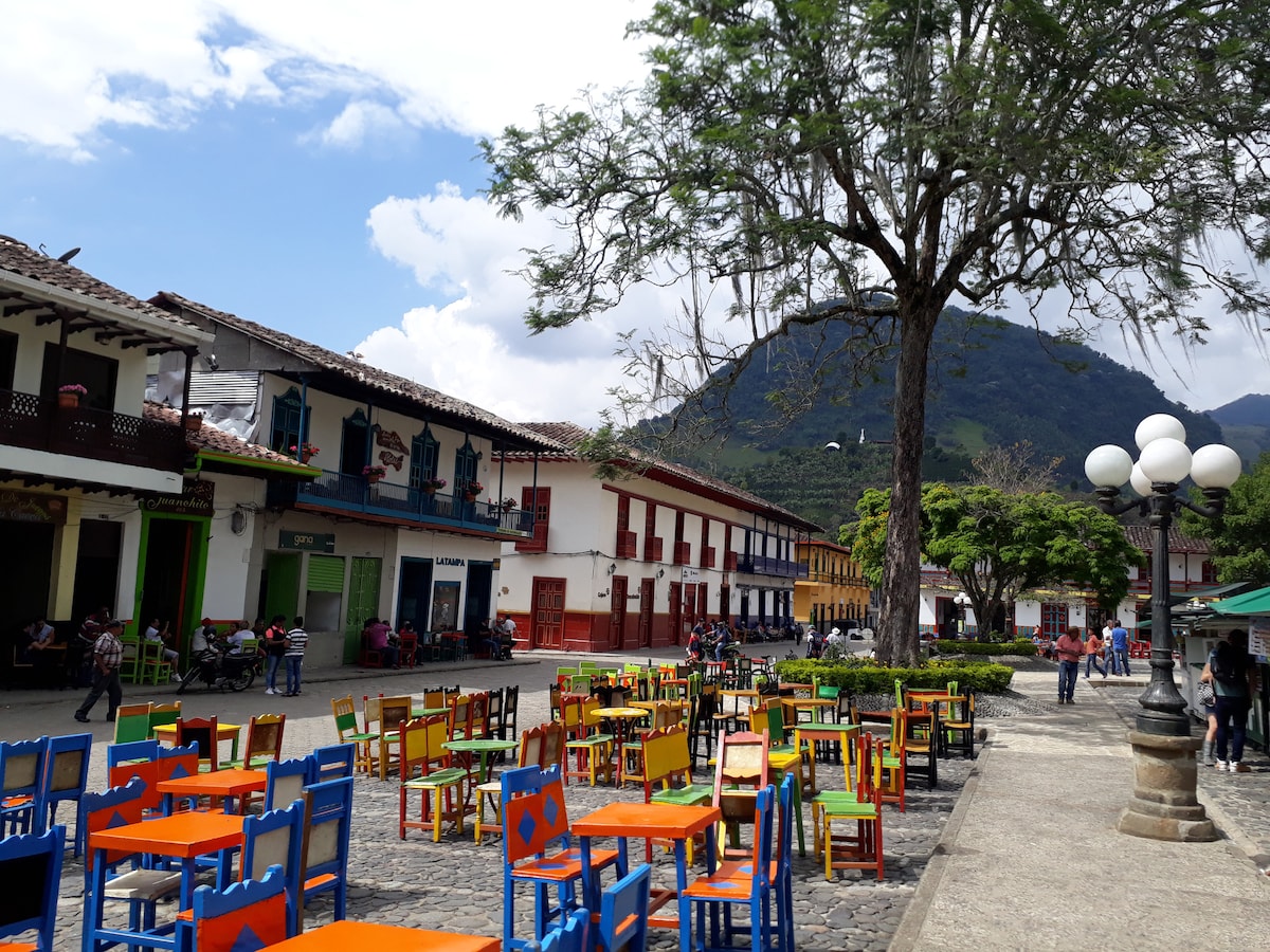 A vibrant square is filled with colorful wooden chairs and tables, set against a backdrop of traditional buildings and a mountainous landscape. Lush trees dot the area, providing shade, while people can be seen enjoying the open space.