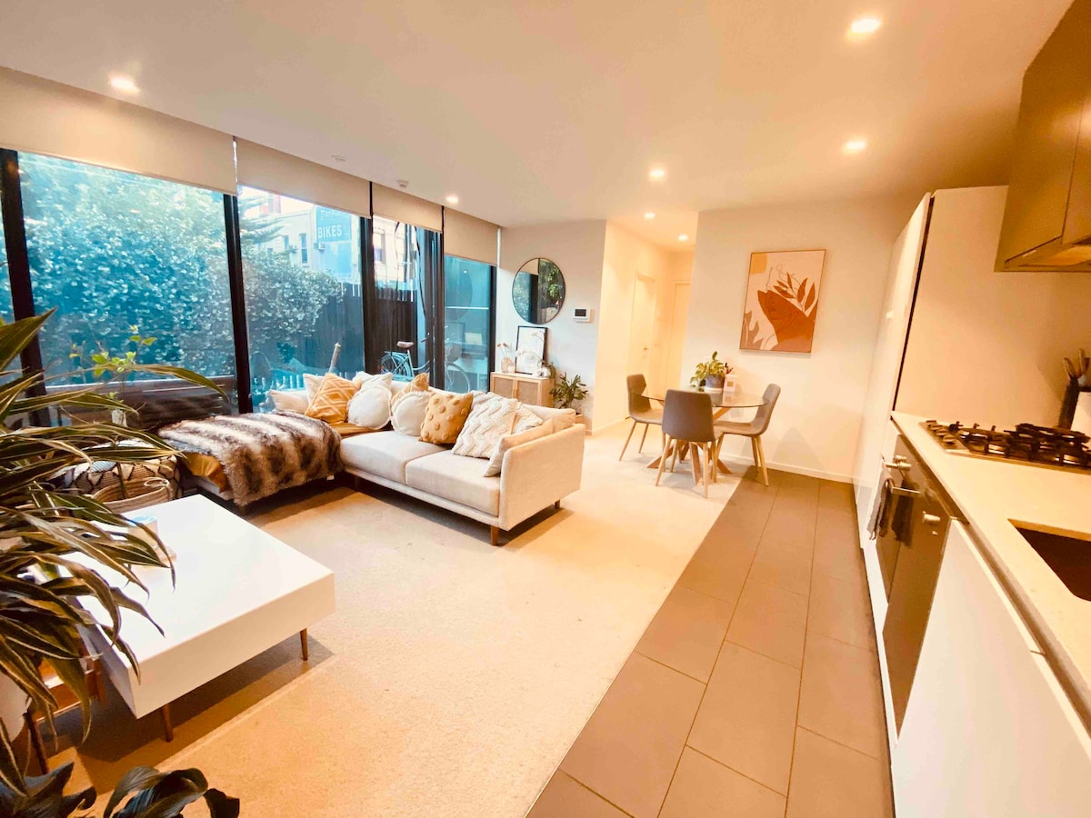 An open-plan living area is presented, featuring a modern sectional sofa adorned with throw blankets. A dining table for four is visible, along with a sleek kitchen equipped with dark cabinetry. Ample natural light illuminates the space through large windows framed by greenery.