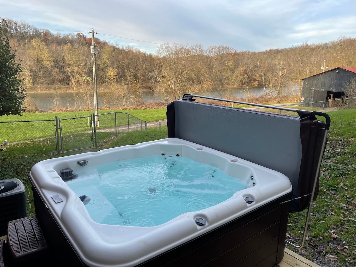 A hot tub is positioned on a deck, featuring a cover partially opened. It overlooks a scenic view of the Kentucky River and surrounding foliage, providing a relaxing outdoor space. The serene landscape includes a grassy area and distant trees along the water's edge.