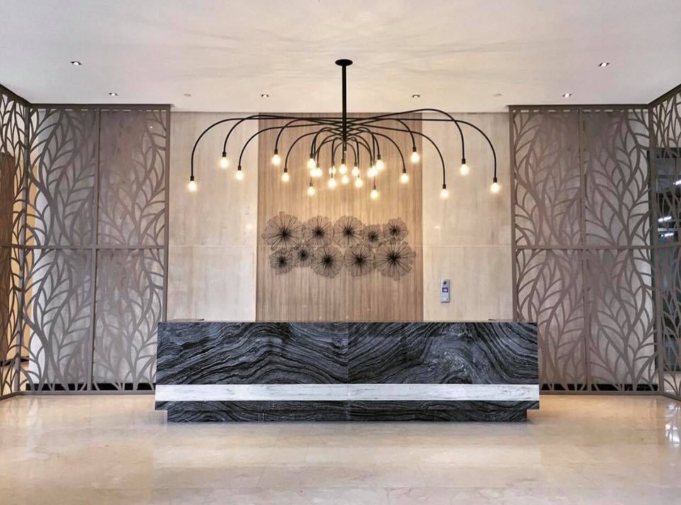 The lobby area showcases a modern reception desk made of dark stone with a contrasting horizontal marble stripe. Behind it, a decorative wall features a floral design. Unique pendant lights hang from the ceiling, creating an inviting focal point.