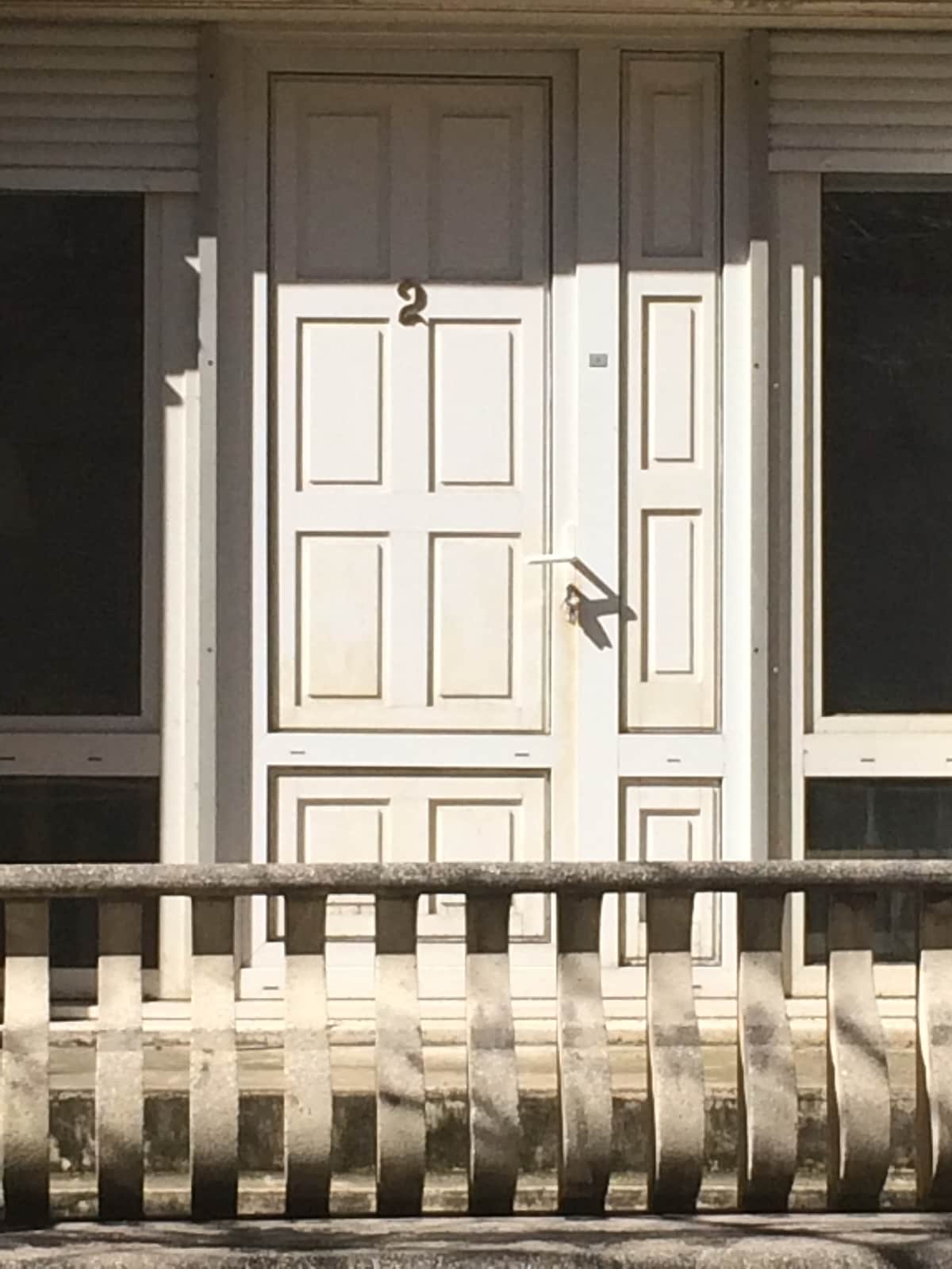 A white door with a large number '2' displayed prominently is framed by a simple guard rail. The minimalist design features a rectangular panel layout and a handle that is positioned to the side. Natural light highlights the textures of the door and surrounding area.