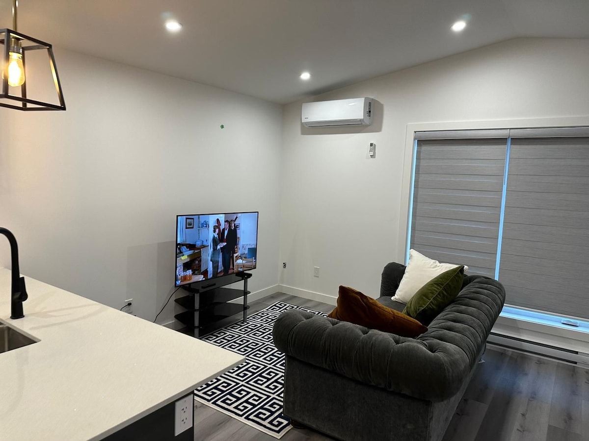 A modern living area showcases a gray sectional sofa adorned with green and white cushions. A wall-mounted television displays an image, while a geometric-patterned rug adds visual interest. Soft lighting highlights the contemporary design, contributing to a relaxed atmosphere.