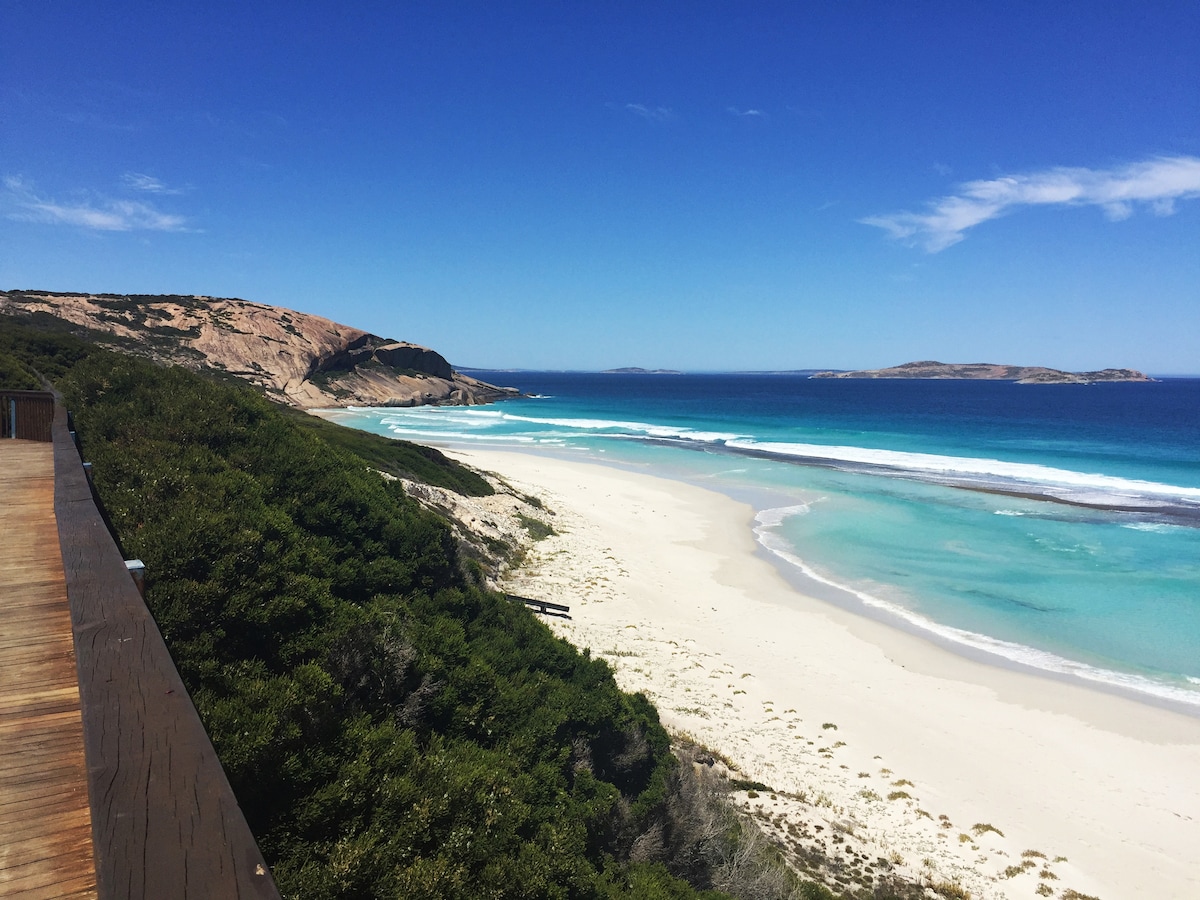 A sweeping view of a serene beach is captured, featuring soft white sands and calm turquoise waters. Rolling hills and a lush green landscape frame the shoreline, while a clear blue sky enhances the tranquil scene, creating an inviting atmosphere for relaxation.
