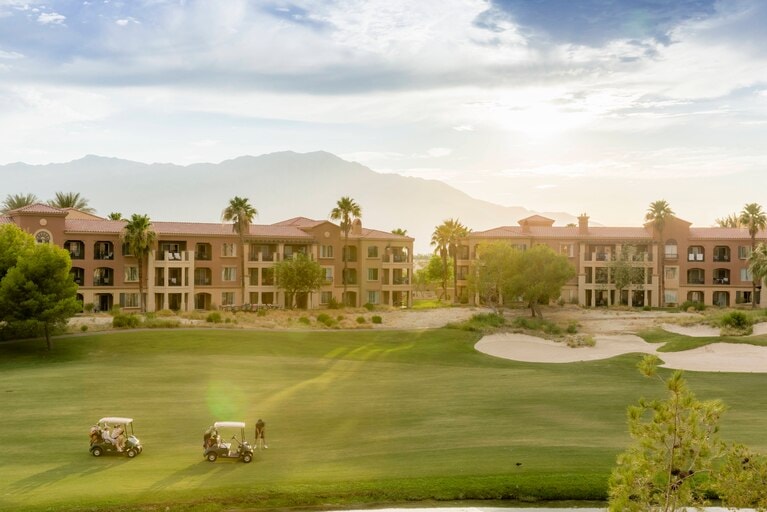 The image features a lush green golf course with two golf carts in motion. Surrounding the course are resort buildings adorned with palm trees, set against a backdrop of mountains and a bright sky with soft clouds. Natural light creates a serene atmosphere.