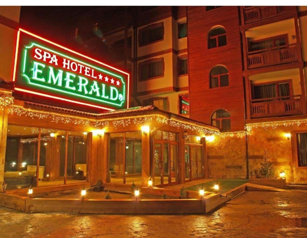 The entrance of Spa Hotel Emerald is illuminated by bright neon signage. Warm lighting highlights the wooden architecture and outdoor pathways, which are lined with subtle garden features. The inviting atmosphere is enhanced by decorative lights along the edges of the building.