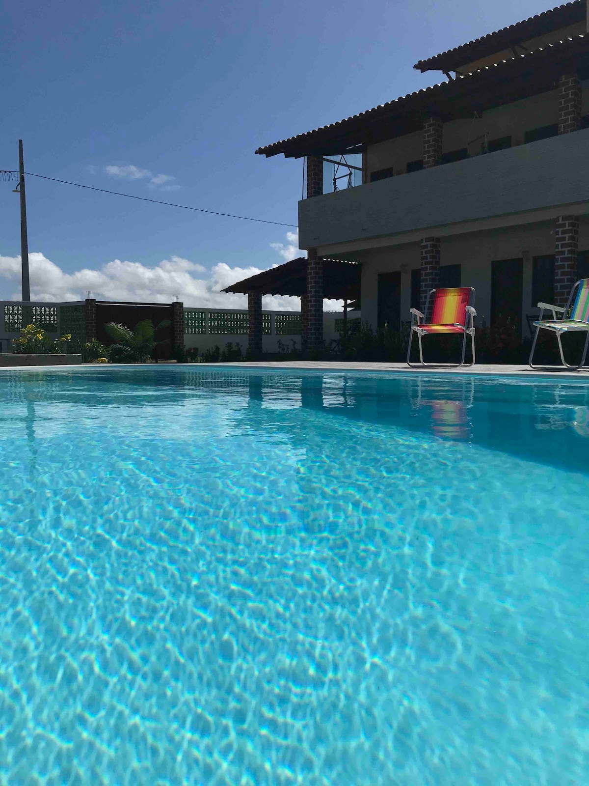 A serene swimming pool is shown with clear blue water shimmering in the sunlight. Two colorful lounge chairs sit near the water’s edge, with gentle ripples reflecting the bright sky and surrounding greenery. A two-story building is visible in the background.