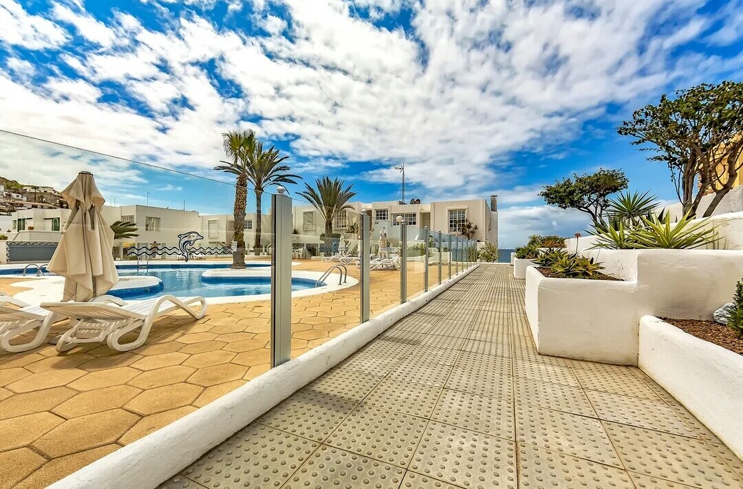 A well-maintained outdoor area features a large pool surrounded by sun loungers and palm trees. The pool is partially shaded by a nearby umbrella. A walkway made of hexagonal tiles leads towards the pool area, offering a clear view of the vibrant blue sky.